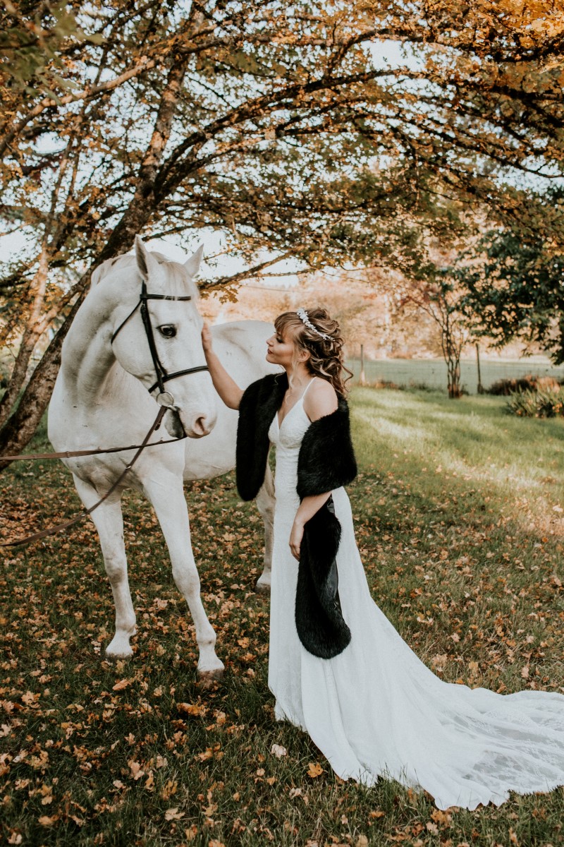 Bride in white lace gown and brown fur stole with white horse Vancouver Island Wedding Magazine