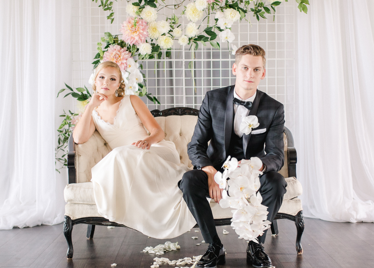 Groom in grey plaid suit holds bouquet of white orchids while bride reclines beside him on cream vintage settee