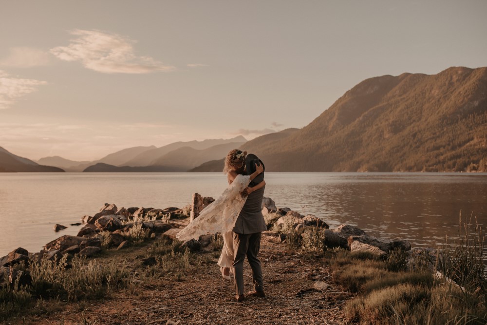 Golden Hour Groom carrying bride by lake mountains in background West coast Weddings Magazine