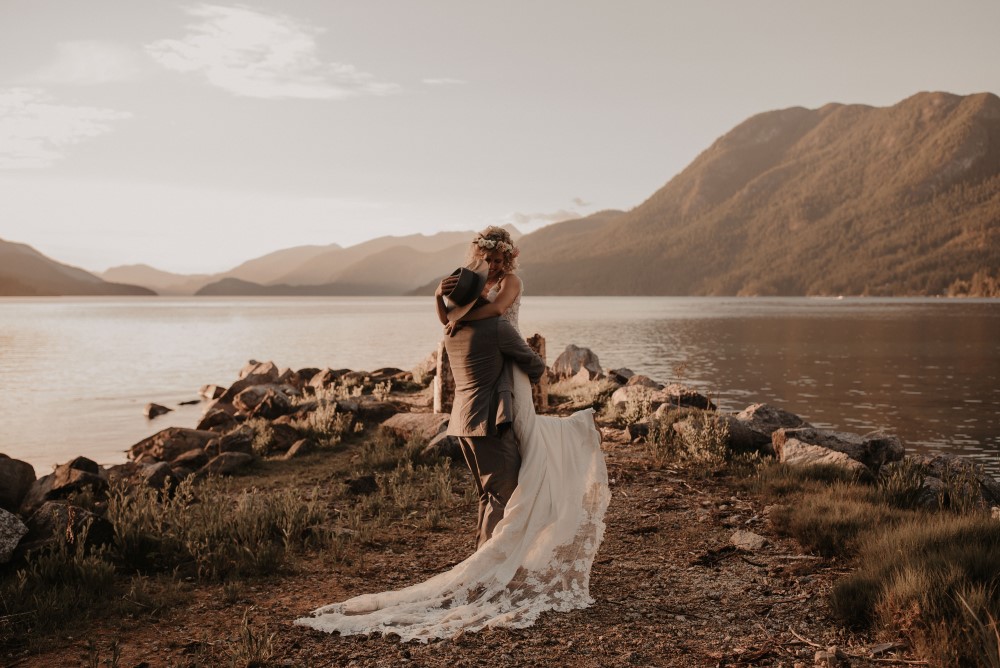 Golden Hour Groom picking up bride with arms around his neck by the lake with mountains Sunshine Coast