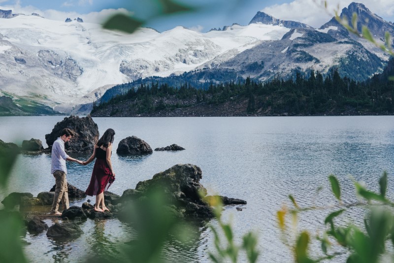 Naturally Romantic Engagement Couple walks on rocks out into BC Lake with mountains behind