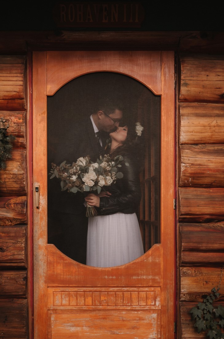 Wedding couple kiss through screen door of rustic cabin
