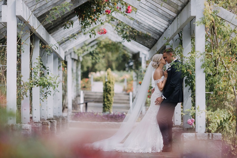 Stanley Park Wedding couplelean against pavilion beam and kiss with roses around them