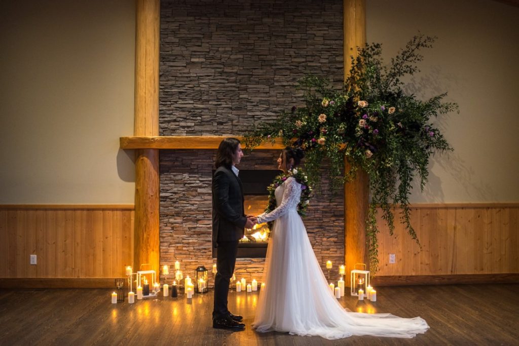 Newlyweds stand in front of large floral arrangement on fireplace mantle