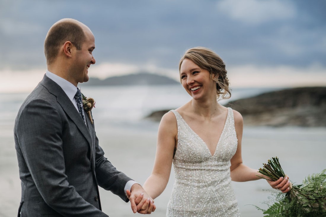 Bride smiles at her groom and holds bouquet on the beaches of Tofino