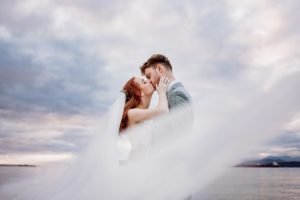 Brides veil swirls around kissing newlyweds with blue sky behind