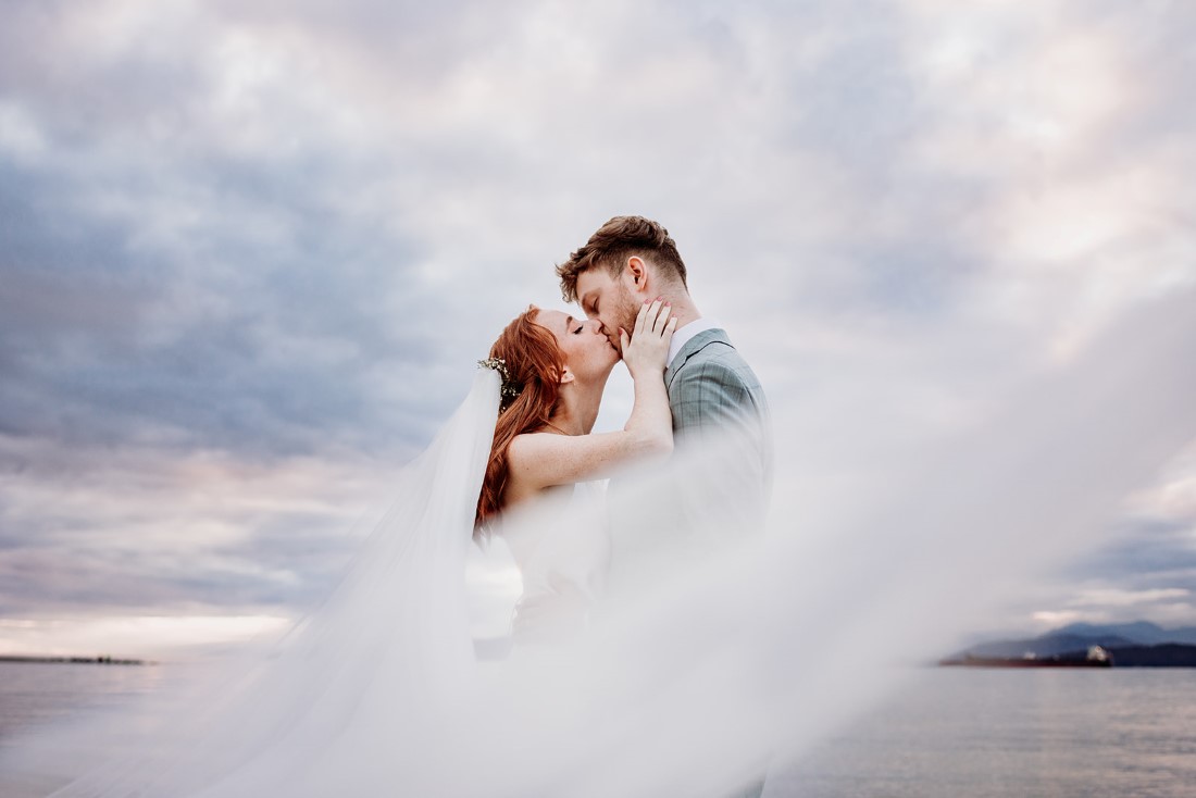 Brides veil swirls around kissing newlyweds with blue sky behind