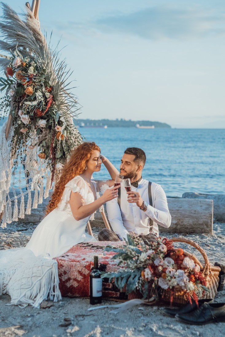 Newlyweds sit by boho teepee with ocean behind