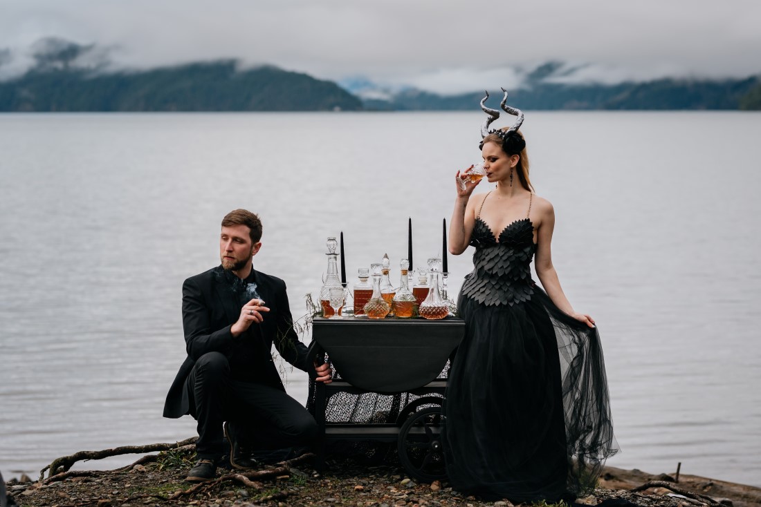 Newlyweds in black gown and suit drink whisky shots in front of Harrison Lake