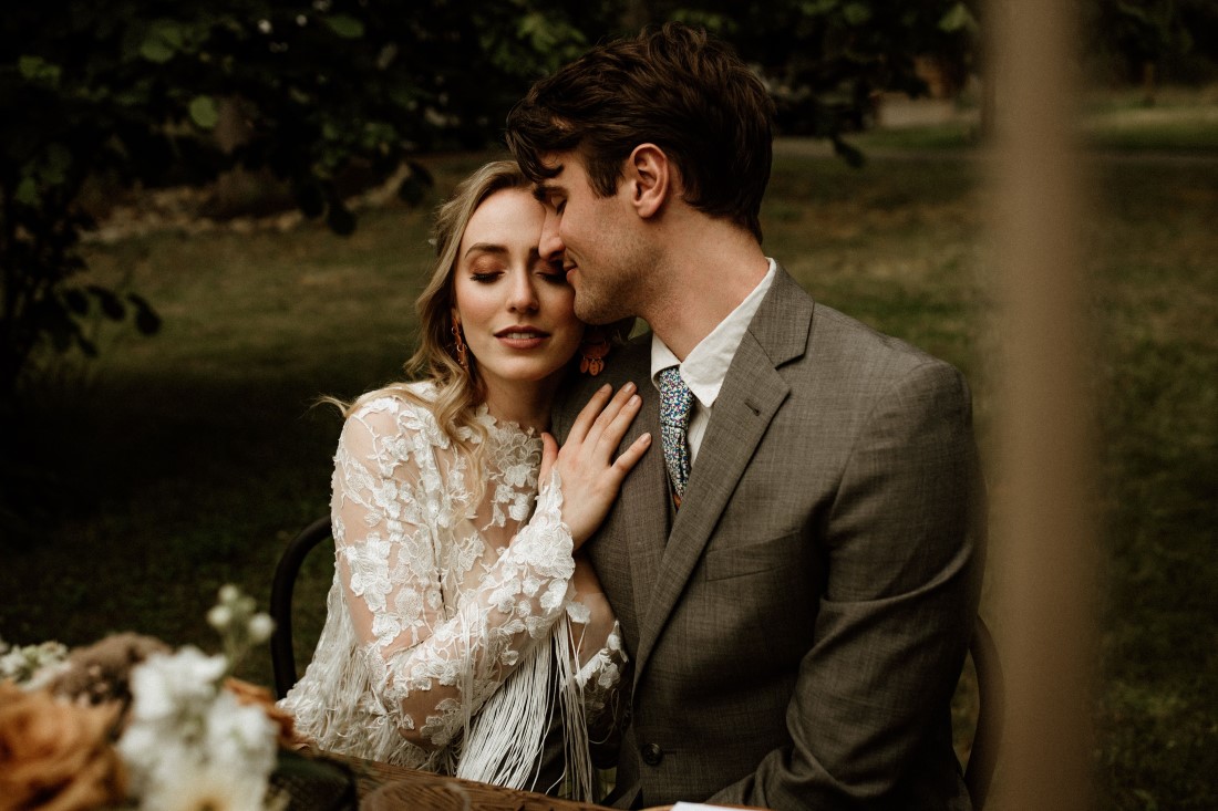 Rustic Romance couple kisses while seated at their outdoor reception table