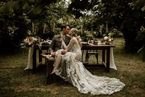Bride and Groom sit at outdoor reception table with lace train by Secret Waters Photo