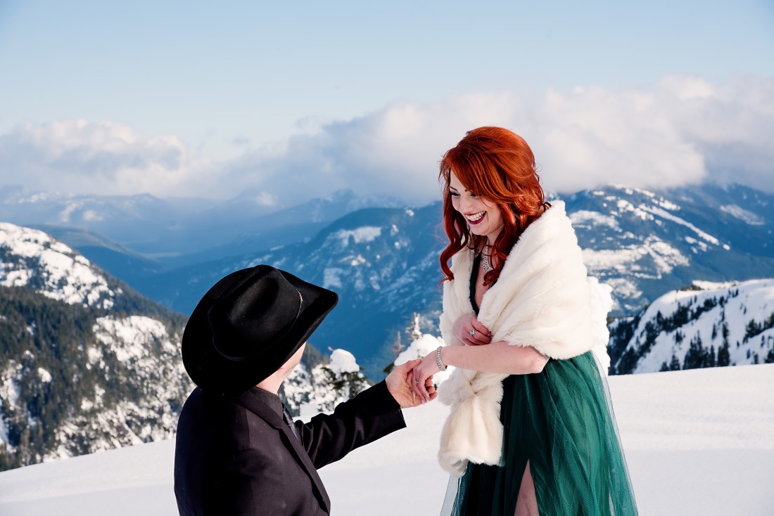 Groom in black cowboy hat down on one knee to propose on top of a mountain