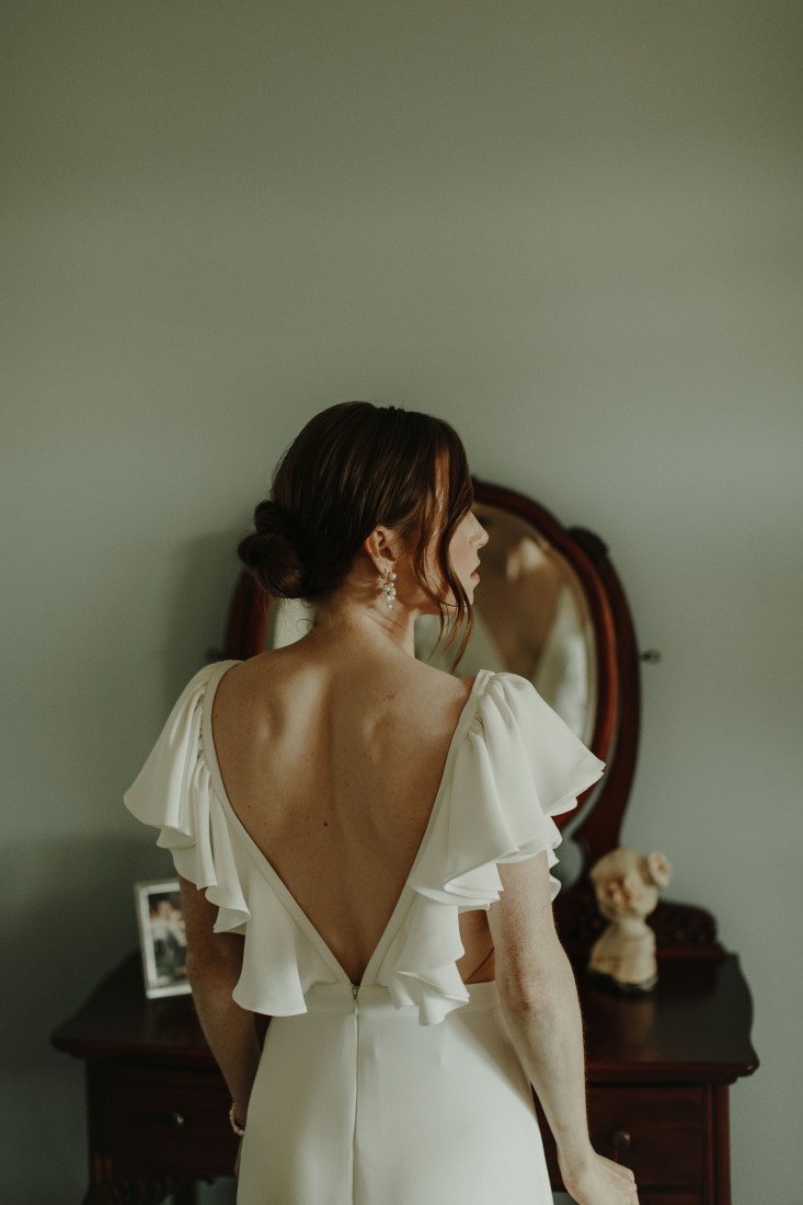Bride with low back lace gown looks in mirror in Vancouver