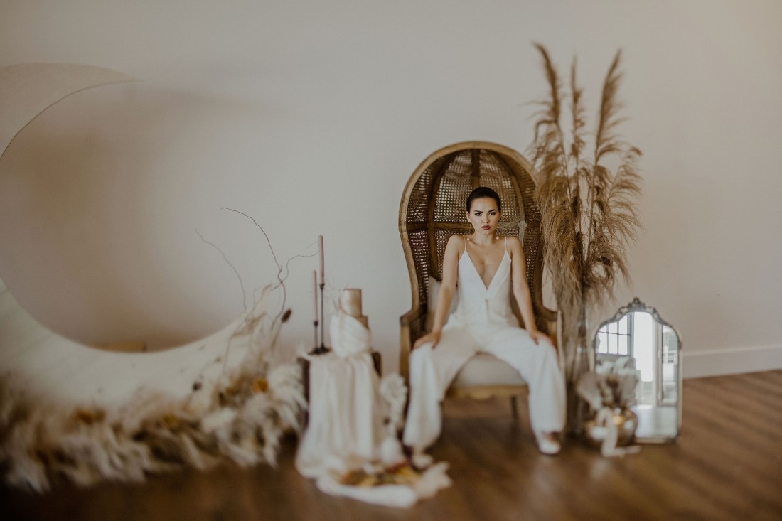 Bride in pantsuit on wicker chair surrounded by boho decor
