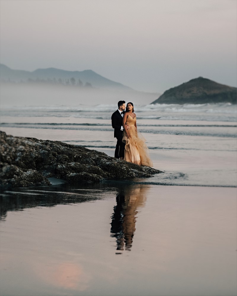 Newlyweds stand on black rock beside Vancouver Island ocean with mist behind
