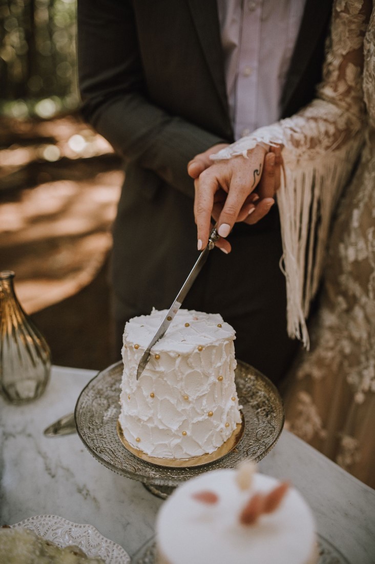 Boho bride with fringed sleeves cuts into white wedding mini cake