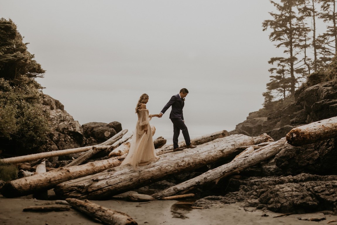 Ethereal Elopement Along Tofino Coastline Couple walks along driftwood on beach