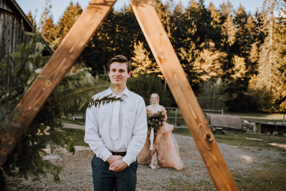 groom stands in front of wood backdrop as bride comes up for first look