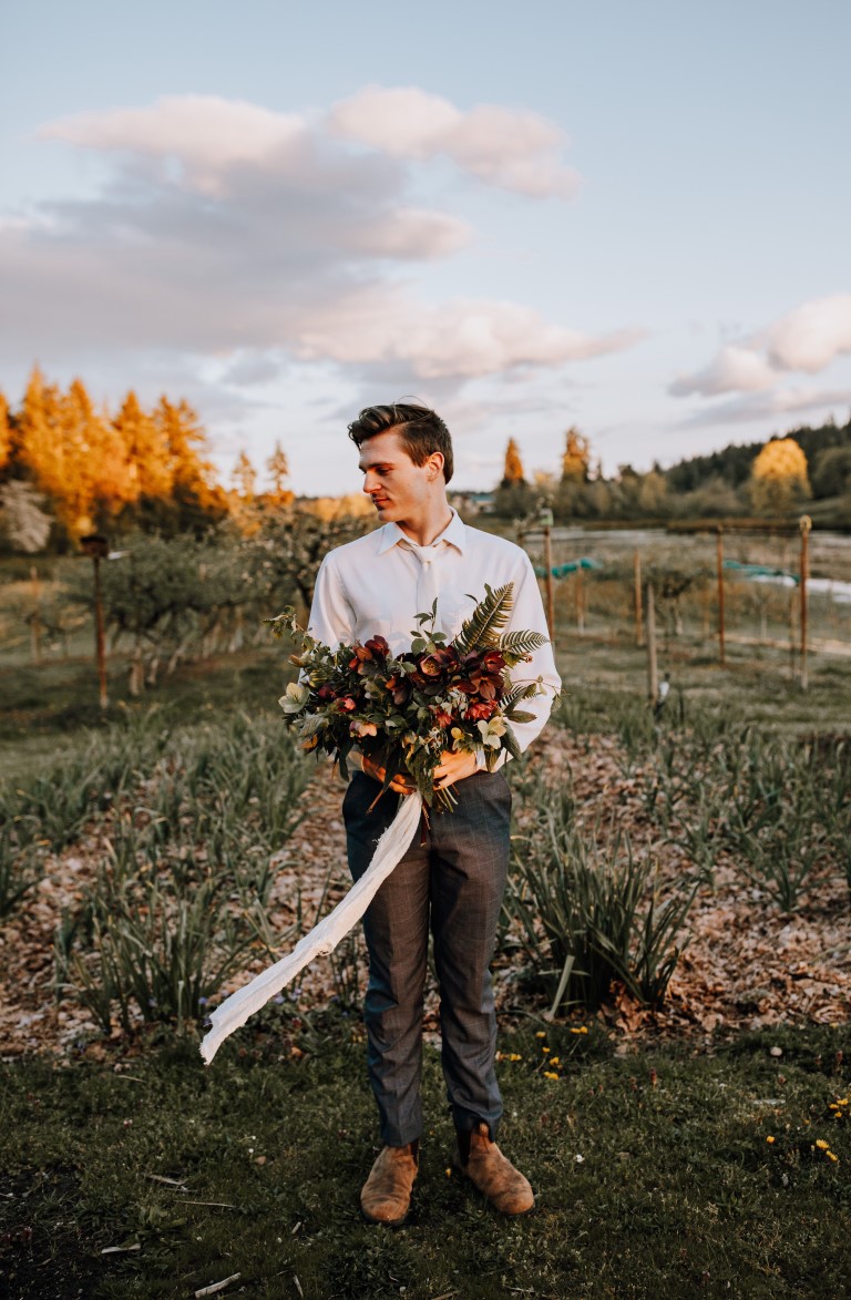 groom stands in farm field holding bouquet of hellebores, ferns, ivy and ribbon