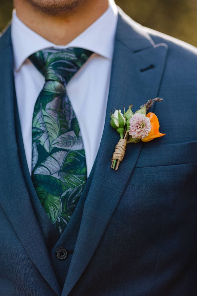 groom wears blue suit and boutonniere with orange rose , white rose bud sprig of purple lavender and pinkish flower