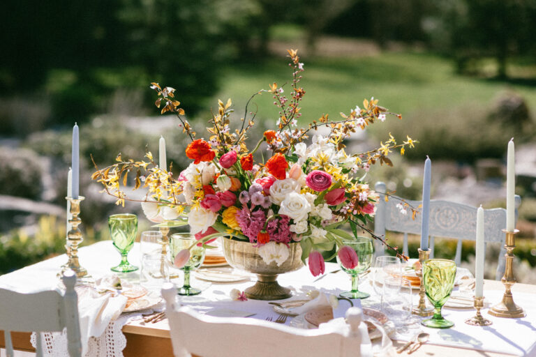 large beautiful floral centerpiece with purple coral, orange, white & pink flowers and greenery. Green glassware and white and blue tapered candles