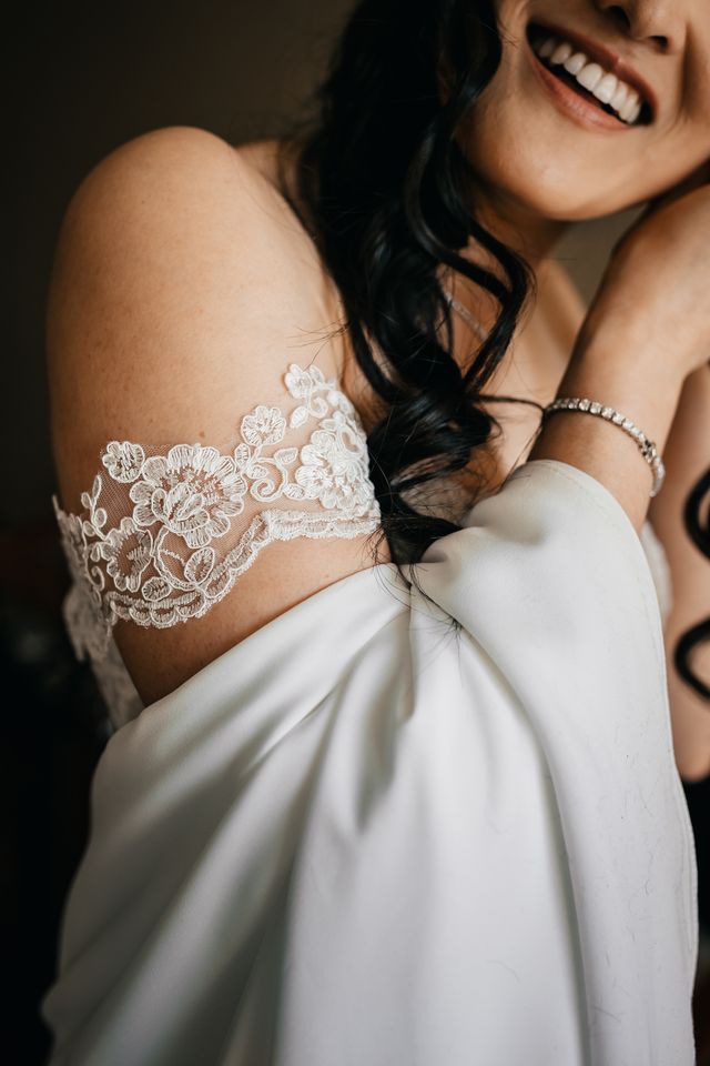 smiling bride puts in her earring while showing her gowns off- the-shoulder lace strap