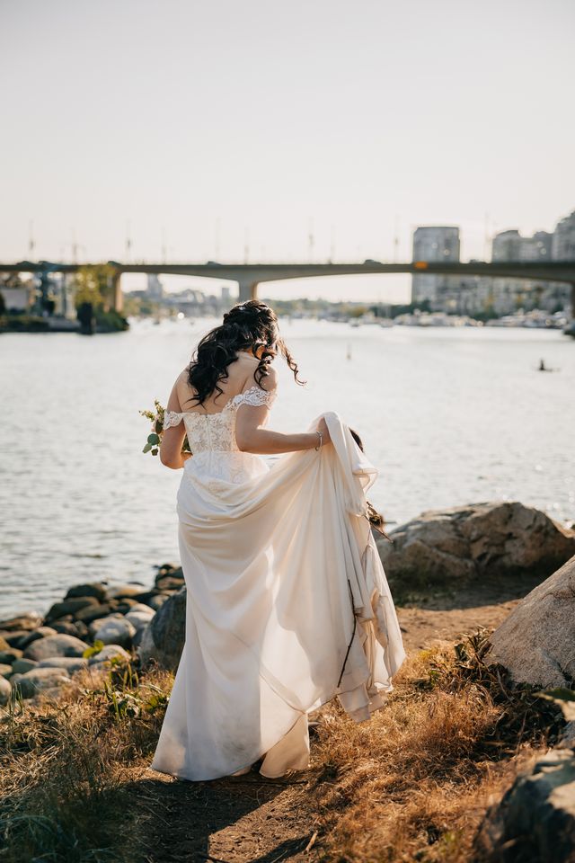 bride holding her dress up on one side and floral bouquet on other ,walks on sand area to seaside