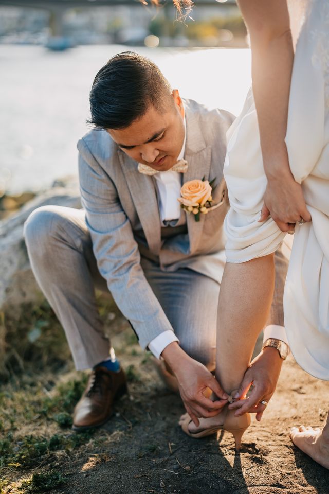 groom helps bride with her shoes on the sand