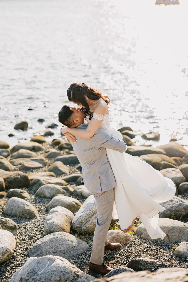 groom lifts bride up at the rocky beach with glistening ocean water in the background