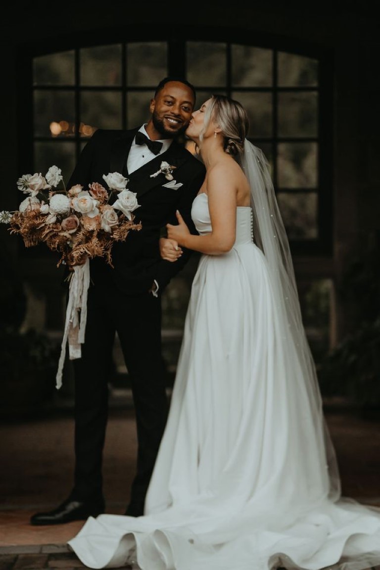 bride kisses her man on the cheek as he holds her large bridal bouquet