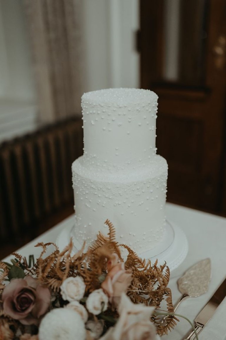 simple white 2 tier wedding cake with pearl details, silver cake server and wedding florals