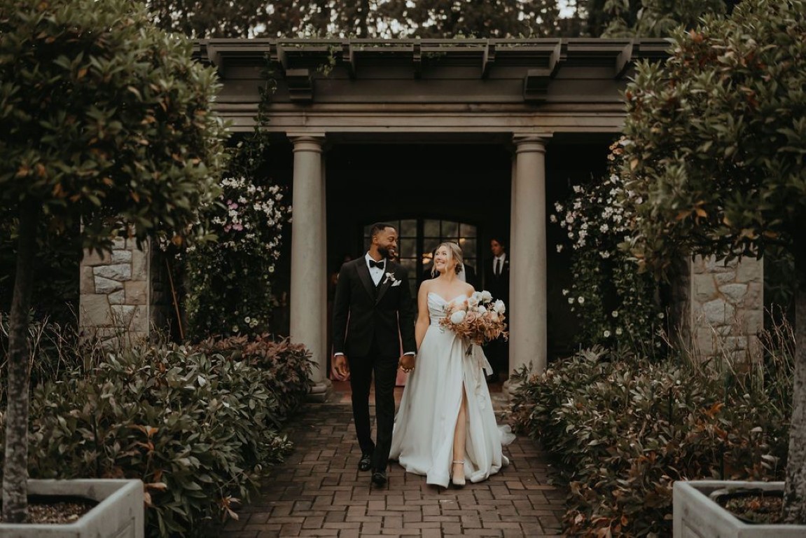 modern bride wears a high slit gown as she walks hand in hand with groom on brick pathway