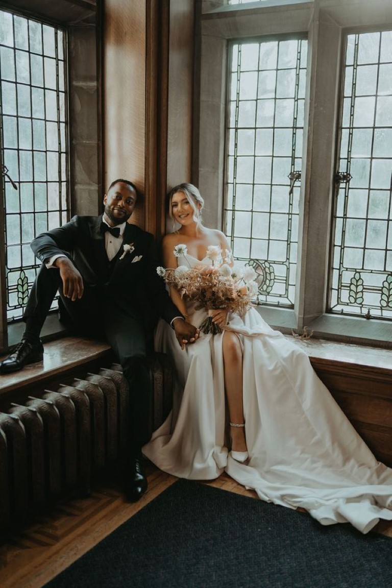 bride and groom sit in corner on wooden window sills with stained glass windows at the castle