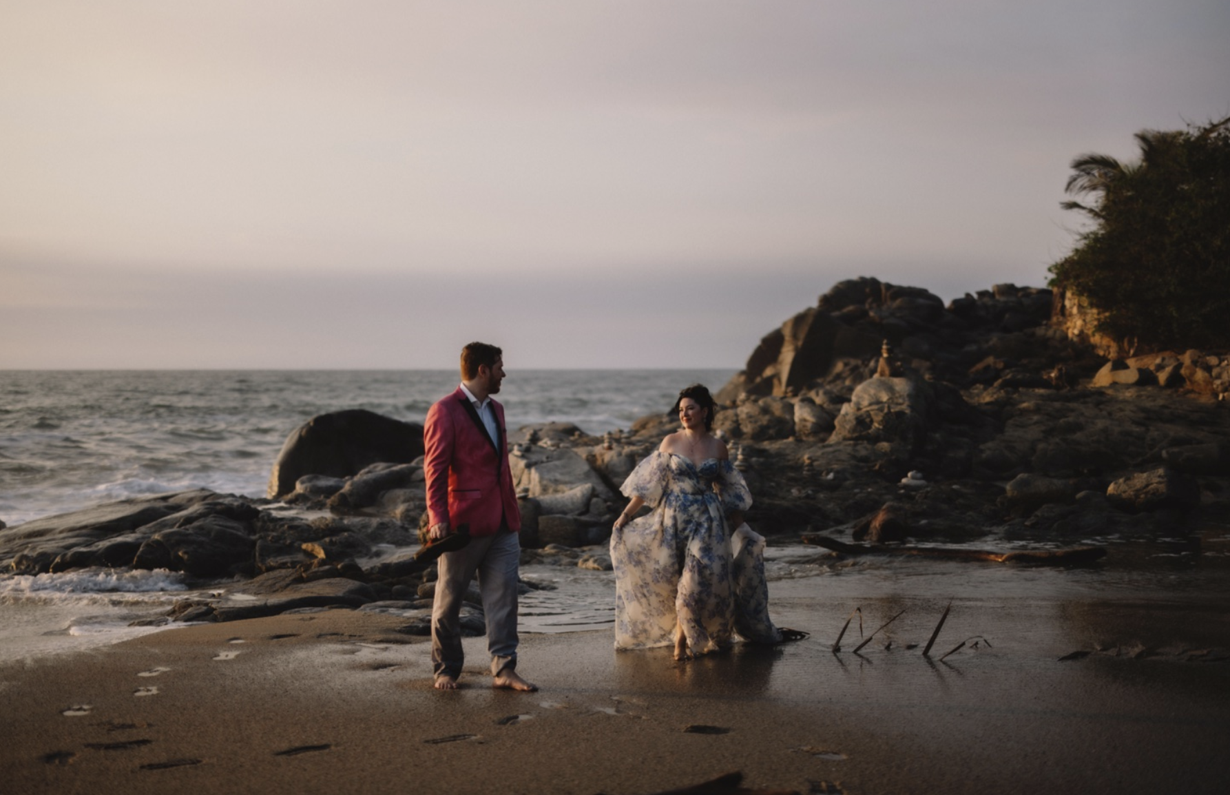 Groom in red suit coat and bride in floral gown walk on the beach by Gabe McClintock