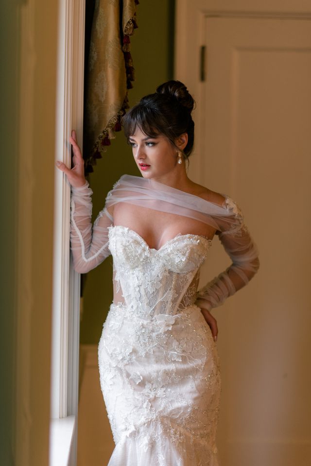 modern bride looks out window dressed in her lace strapless gown with tulle long sleeve detail