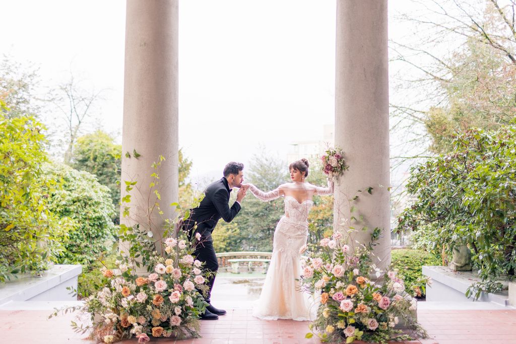 groom kisses brides hand as she leans on large pillar on the manors veranda