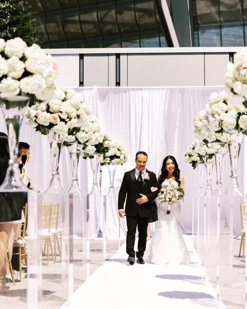 Father walks bride down aisle surrounded by white flowers PROLINE