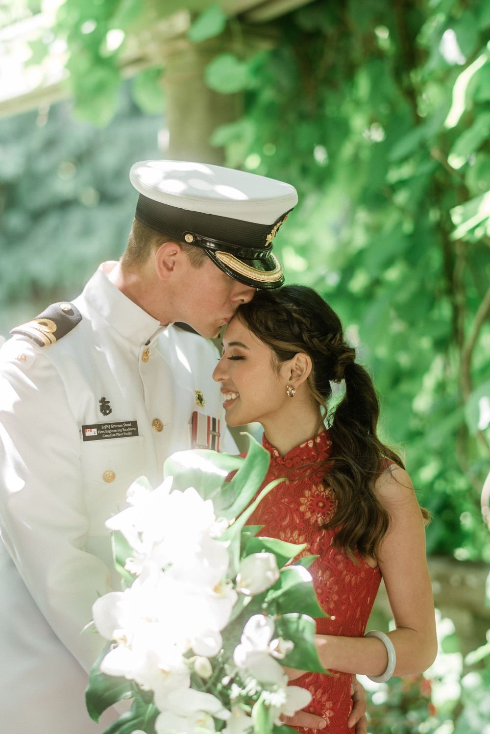 Groom in dress whites and hat kisses bride in red lace gown