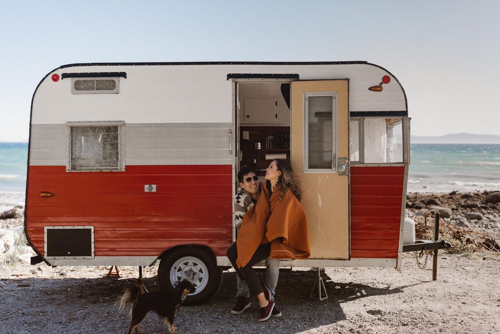 Engaged Couple and black dog in door of Orange Retro Trailer in desert area