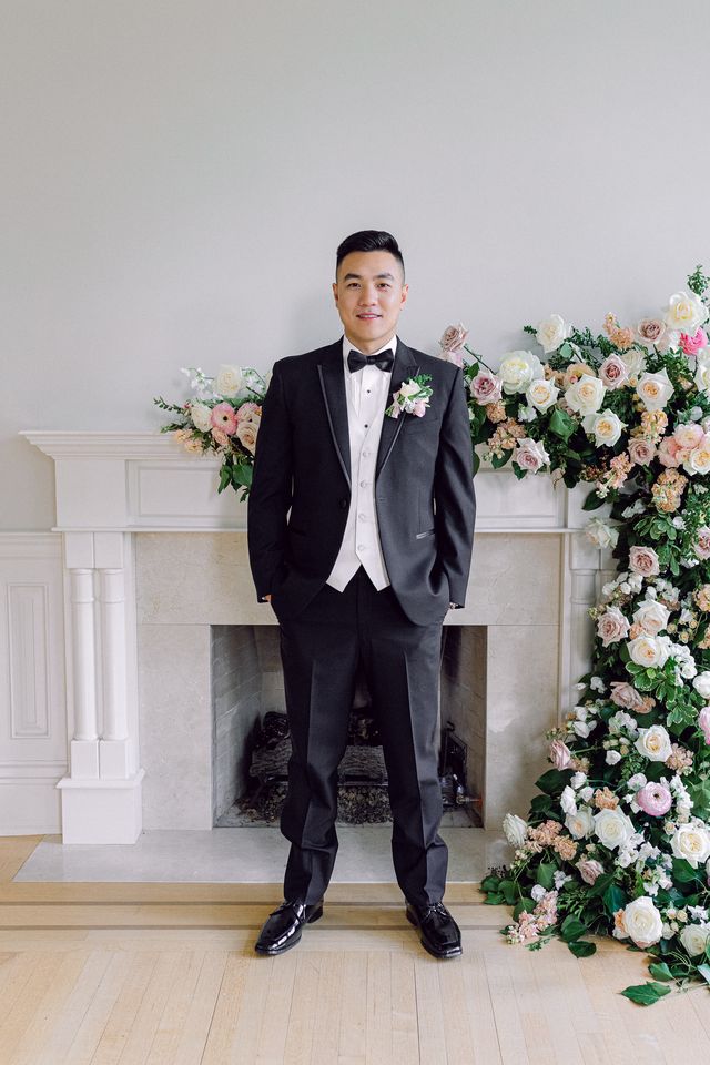 Groom stands in front of floral design over fireplace mantle