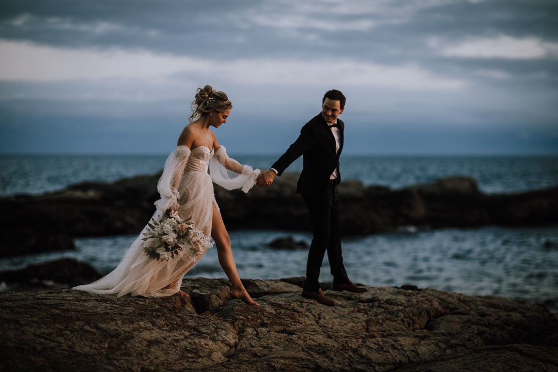 Bride and groom walk across beach with blue sunset behind